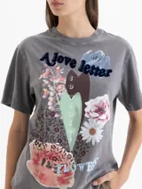 Remera gris con efecto lavado, de corte holgado y cuello redondo. Presenta una estampa frontal con motivos florales, un corazón central y la inscripción 'A love letter to winter flowers'. Cuenta con tajos laterales en el ruedo.