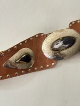 Cinto tipo faja de cuero color beige, con apliques metálicos plateados de forma ovalada y de gota, decorados con piedras oscuras y tachas metálicas a lo largo de toda la pieza.