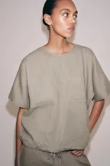 Top de lino de color beige, con cuello redondo, manga corta, bolsillos frontales aplicados y bajo ajustable con cordones. Cuenta con abertura en la espalda y cierre de botón.