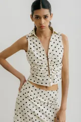 Chaleco blanco con estampado de lunares negros, cuello camisero y cierre frontal con broches metálicos.