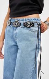 Cinto de cuero negro con diseño de piezas circulares unidas por argollas metálicas, decorado con tachas plateadas y tiras largas de cuero colgantes en el lateral.