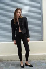 Blazer corta de estilo militar en color negro, con cuello redondo, manga larga y hombreras marcadas. Presenta un diseño frontal con bordados decorativos a tono y botones dorados, cierre con gancho metálico y forro interior.