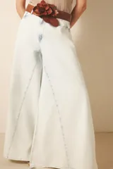 Jean de tiro alto y corte ultra ancho, confeccionado en denim rígido con lavado bleach que le otorga un tono casi blanco. Presenta recortes estratégicos en las piernas y el tiro, brindando una silueta con gran vuelo y movimiento. Incluye cierre y botón frontal, además de forrería interna estampada.