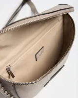 Riñonera de cuerina color gris taupe, con diseño semicircular, cierre superior y detalles de tachas metálicas en el borde inferior. Incluye correa ajustable y placa metálica con la inscripción de la marca.