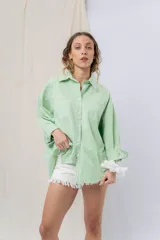 Camisa verde pastel de algodón, corte oversize, con cuello camisero, cierre con botones, mangas largas con puños con volados de encaje blanco, dos bolsillos frontales con solapa y ruedo irregular. 