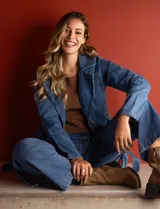 Chaqueta larga de jean azul con cuello con solapa, abertura central con botones, bolsillos delanteros, cintura con pasadores y lazo de ajuste, y puños con botón.