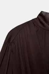 Blusa corta marrón de tejido fluido y semitransparente, con cuello subido, diseño cruzado drapeado en el frente y mangas largas abullonadas con puños ajustados.