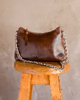 Cartera tipo media luna de cuero natural color marrón, con cierre superior y correa de cadena metálica plateada. Presenta el logo de la marca grabado en el frente.