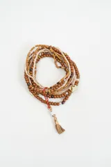 Brazalete multicapa compuesto por varias hileras de cuentas en tonos tierra, beige y dorado. Incluye un dije de corazón dorado y un colgante con borla de gamuza y detalle de piedra blanca.