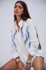 Camisa blanca de manga larga con cuello clásico y botones frontales. Presenta un diseño de ondas en tonos azules con lentejuelas y pequeños apliques metálicos en el frente.