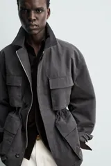 Cazadora relaxed fit de hombre, confeccionada en tejido técnico con algodón de color negro. Cuenta con cuello camisero, manga larga y bolsillos frontales de plastrón con solapa. Presenta cintura ajustable con cordón interior y cierre frontal con cremallera oculta por solapa con botones a presión.