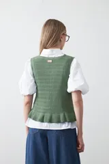 Chaleco tejido color verde musgo, con trama texturada, cuello a la base y cierre frontal con lazos. Presenta un ruedo con volado.