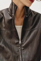 Chaqueta estilo bomber corta de efecto piel en color marrón oscuro, con cuello subido, cierre frontal con cremallera y cintura elástica fruncida.