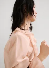 Blusa de seda color rosa pálido con volados frontales, cuello redondo y mangas largas con puños elásticos.