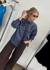 Camisa de denim azul con diseño en canesú y mangas anchas. Presenta bordados florales oscuros en el frente y detalles de ribetes oscuros en cuello y costuras.