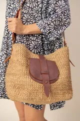 Bolso tipo bandolera confeccionado en rafia natural de color beige, con detalles de cuero marrón en la correa ajustable, los laterales y un bolsillo frontal con solapa y cierre de tira pasante.