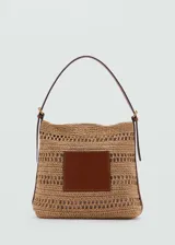 Bolso tipo hobo confeccionado en tejido de fibra natural con diseño calado. Presenta un parche central de cuero sintético, asa de hombro ajustable con hebillas metálicas y cierre magnético.