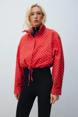 Chaqueta roja con estampado de lunares blancos, cuello subido, manga larga con puños plisados, bolsillos delanteros, bajo ajustable con cordón y cierre frontal con cremallera.