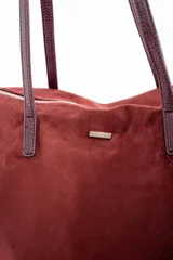 Bolso tipo tote de gran tamaño en color bordó, confeccionado en material sintético con textura de gamuza. Cuenta con cierre superior, correas largas de hombro en tono marrón oscuro y una placa metálica decorativa en el frente.
