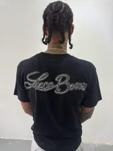 Remera de algodón color negro con cuello redondo y estampa gráfica blanca en el pecho. Presenta un corte regular y una pequeña etiqueta decorativa en la manga.