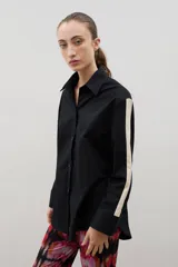 Camisa de corte holgado y largo medio, color negro con detalle de franja lateral en color crema a lo largo de las mangas. Presenta cuello clásico, cierre frontal con botones y tajos laterales en el ruedo.