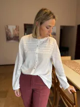 Camisa de lino color blanco con cuello redondo, cierre frontal de botones y detalle de alforzas y encaje en el pecho. Presenta mangas largas con puños abotonados y un corte recto.