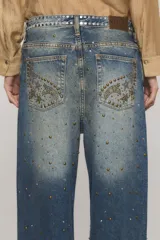 Jeans de tiro medio y calce holgado, color azul con lavado desgastado. Presenta tachas metálicas aplicadas en la parte frontal formando patrones decorativos y en la pretina.