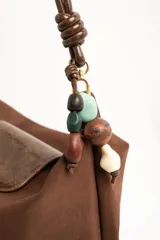 Cartera tipo shopper color marrón, con solapa frontal decorativa en tono más oscuro y tiras con nudos. Incluye herrajes dorados y un charm colgante con cuentas de madera.