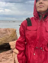 Piloto impermeable de color rojo con detalles de volados verdes en los hombros. Presenta capucha integrada, cintura ajustable con cordón, mangas con volumen y puños elastizados.