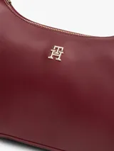 Cartera tipo hobo de Tommy Hilfiger en color bordó, confeccionada en poliuretano con acabado liso. Cuenta con compartimento principal con cierre de cremallera, bolsillo interior con cierre, asa superior y correa ajustable y extraíble. Presenta el logo metálico de la marca en relieve en la parte delantera y detalles en herrajes dorados.