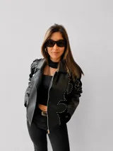 Chaqueta tipo biker de eco cuero negro con diseño estructurado. Presenta apliques de tachas metálicas en formas orgánicas sobre paneles de gamuza sintética, cierre frontal metálico y bolsillos laterales con cierre.