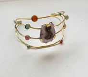 Brazalete rígido dorado con tres hileras de metal y piedras naturales engarzadas de diferentes colores. Presenta una piedra de amatista en el centro.