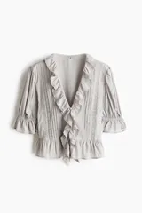 Blusa color gris claro en tejido de mezcla de viscosa y algodón, con escote en V pronunciado, volados frontales, alforzas, inserciones de encaje y detalles bordados. Cuenta con mangas abullonadas al codo con puños elásticos de volado y volado ancho en el bajo.