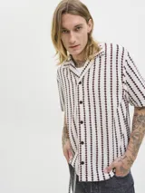 Camisa resort de manga corta color beige, con patrón geométrico de rayas verticales en tonos bordó, cuello camisero, cierre frontal de botones y calce relajado.