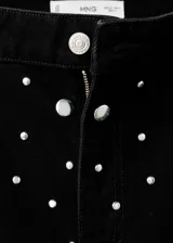 Jeans rectos de tiro medio en tejido de algodón vaquero color negro, con diseño de cinco bolsillos, trabillas para cinturón y cierre frontal con botón y cremallera. Presenta aplicaciones de tachuelas brillantes a lo largo de las piernas.