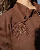Chaqueta de estilo utilitario color marrón, con cierre frontal completo, cuello camisero, dos bolsillos frontales tipo parche y un bolsillo pequeño con cierre en el pecho. Presenta un bordado decorativo en el pecho y puños con botones.