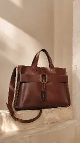 Bolso tipo shopper de cuero marrón, con diseño estructurado, asas superiores y correa larga regulable. Presenta una banda decorativa horizontal con hebilla central y un dije colgante con marca grabada.