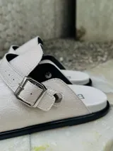 Zueco negro de cuero sintético con textura, estilo cerrado y tira ajustable con hebilla metálica plateada.