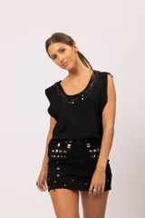 Pollera mini de denim negro con terminación desflecada. Presenta un diseño decorado con múltiples tachas cuadradas plateadas de diversos tamaños distribuidas en la cintura, sobre las solapas de los bolsillos delanteros y en el frente.