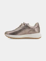 Sneaker de mujer con acabado metalizado en tono dorado, diseño con perforaciones decorativas en los laterales y cordones al tono. Presenta una suela de plataforma blanca con efecto moteado y base de goma color caramelo.