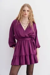 Vestido corto de color violeta, con escote cruzado adornado con ojales metálicos, mangas largas abullonadas con puños elásticos y falda con volado en el ruedo. Presenta un lazo ajustable en la cintura y un sutil diseño de corazones en la tela.
