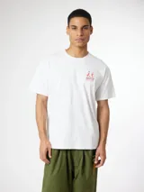 Camiseta blanca de algodón orgánico, con corte oversize, cuello redondo y manguita corta. Presenta un pequeño estampado rojo en el lateral superior izquierdo con la frase 'Go Out And Slide' y caracteres en japonés junto a una ilustración de dos personas cargando una tabla de surf.