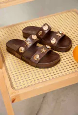 Sandalias planas de color marrón con dos tiras anchas, decoradas con apliques metálicos dorados en forma de caracoles y estrellas de mar.
