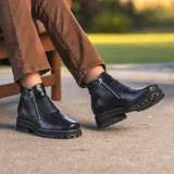 Bota corta de cuero vacuno color negro, con diseño de punta cuadrada y suela de goma tipo track. Presenta doble cierre metálico lateral y una estética robusta y moderna.