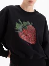 Buzo negro de corte holgado con cuello redondo, mangas largas y puños acanalados. Presenta un estampado central de una frutilla grande con detalles realistas.