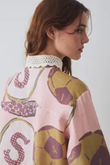 Camisa de lino blanca con estampado de motivos deportivos, cuello y puños tejidos a mano en crochet azul con borde rojo. Presenta cierre frontal con botones y un bolsillo en el pecho.