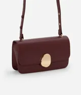 Cartera tipo shoulder bag de tamaño mediano, confeccionada en poliuretano color marrón oscuro. Presenta una solapa frontal con un detalle metálico dorado de forma orgánica y asa ajustable.