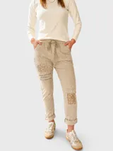 Pantalón de corte relajado color beige, con cintura elástica y cordón ajustable. Presenta parches decorativos con lentejuelas y tachas en los muslos, además de detalles de texto estampado. Posee bolsillos laterales y dobladillo en los tobillos.