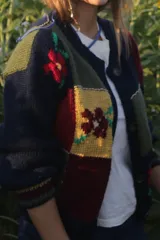 Saco tipo cardigan de tejido de punto con diseño patchwork multicolor, presentando bloques en tonos azul marino, amarillo, rojo y verde con detalles de flores bordadas. Cuenta con cierre frontal de botones y puños acanalados.
