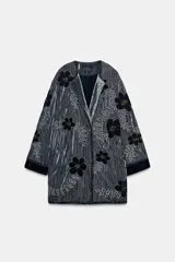Kimono de terciopelo gris con acabado texturizado, cuello redondo y manga larga. Presenta bordados florales en hilo negro y metalizado, bolsillos delanteros y cierre frontal.